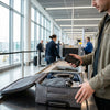 Reiziger houdt een powerbank en boardingpass vast bij airport security, met open handbagage en luchthavenachtergrond in zacht daglicht.